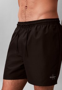 Άνδρας που φοράει μαύρα μαγιό σορτς Calvin Klein με σούρα στη μέση, εμφανιζόμενος από τη μέση μέχρι το μέσο του μηρού, με γκρίζο φόντο.
