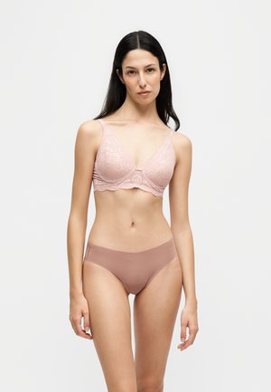 Kvinde med langt mørkt hår iført lyserød blonde-bh og matchende mauve sømløst undertøj, stående mod en ensfarvet hvid baggrund.