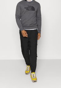 Graues Sweatshirt mit schwarzem Logo, schwarze Hose mit verstellbaren Bündchen und gelbe Turnschuhe mit grauen und schwarzen Akzenten.