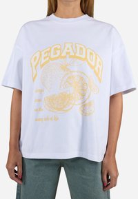 T-shirt blanc surdimensionné avec un imprimé graphique jaune représentant des oranges et le texte "PEGADOR" ainsi que "toujours se concentrer sur le bon côté de la vie."