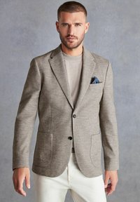 Hellbrauner kariertes Blazer mit zwei vorderen Taschen, schwarzen Knöpfen und einem blauen gemusterten Taschentuch, kombiniert mit einem grauen Pullover und cremefarbenen Hosen.
