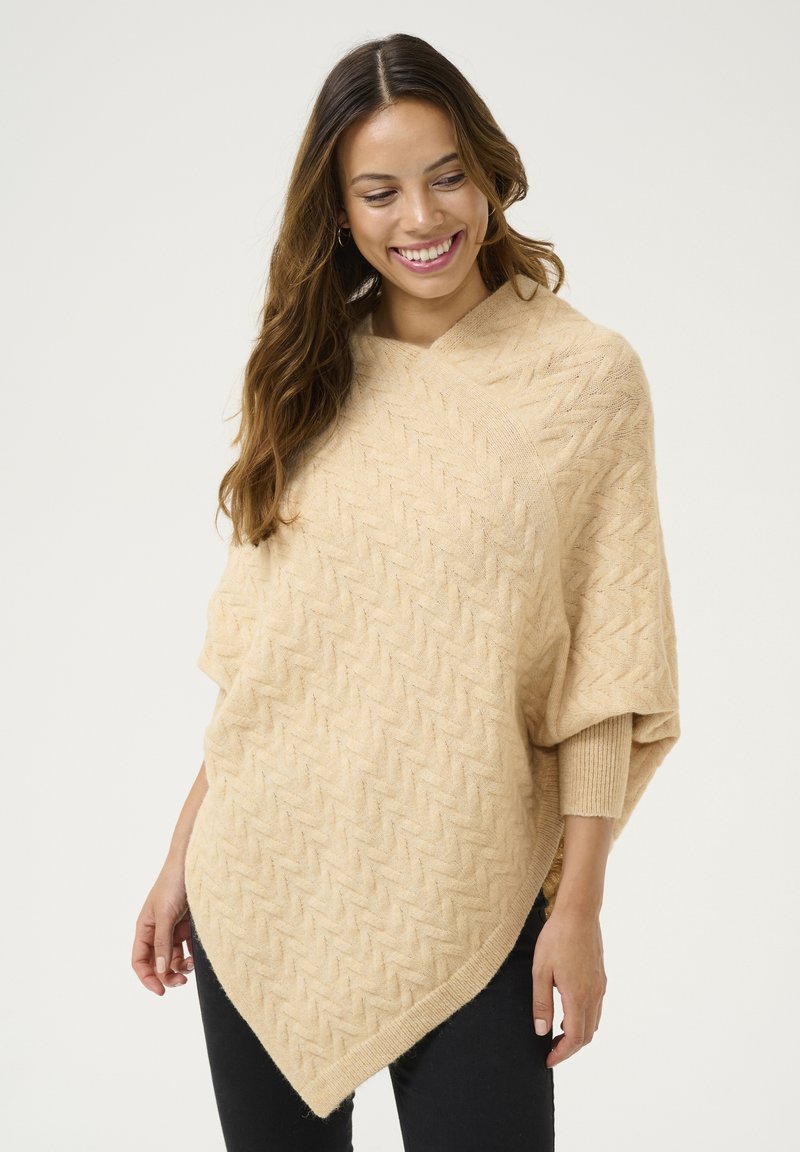 Beige gebreide poncho met een visgraatpatroon, voorzien van een V-hals ontwerp en een puntenzoom. Zachte textuur, ontspannen pasvorm en opgerolde mouwen.
