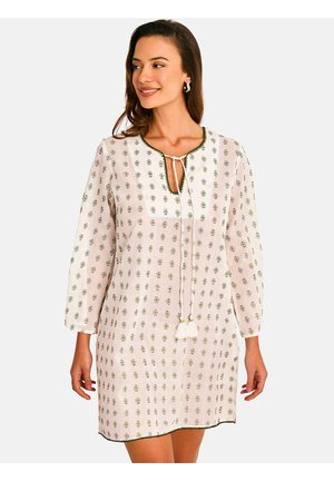 Femme portant une robe blanche avec des motifs floraux verts, un col en V avec des liens, et des manches trois-quarts, souriante et regardant sur le côté.