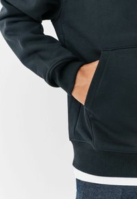 Schwarzer Hoodie mit gerippten Bündchen und Saum, mit einer vorderen Kängurutasche. Glatte Textur und minimalistisches Design. Über einem weißen Hemd getragen.