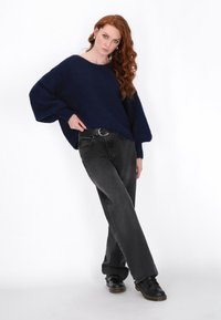 Granatowy, oversize'owy sweter z dzianiny z długimi rękawami, zestawiony z prostymi czarnymi dżinsami i czarnymi butami, stojący na białym tle.