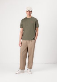 Olivenfarget bomulls-T-skjorte med korte ermer og broderi på brystet, kombinert med beige vide bukser og hvite sneakers. Lys caps på hode.