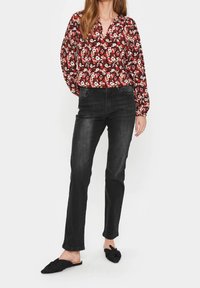 Röd blommig blus med V-ringning, långa ärmar och svarta jeans, tillsammans med svarta knutna slip-on-skor, uppvisad mot en vit bakgrund.