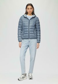 Helle blaue Steppjacke mit Kapuze, ausgestattet mit einem gesteppten Design und zwei Fronttaschen. Kombiniert mit hellblauen Jeans und silbernen Turnschuhen.