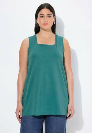 Vrouw met lang donker haar draagt een teal mouwloos topje met vierkante hals en een blauwe spijkerbroek, staand tegen een effen witte achtergrond.