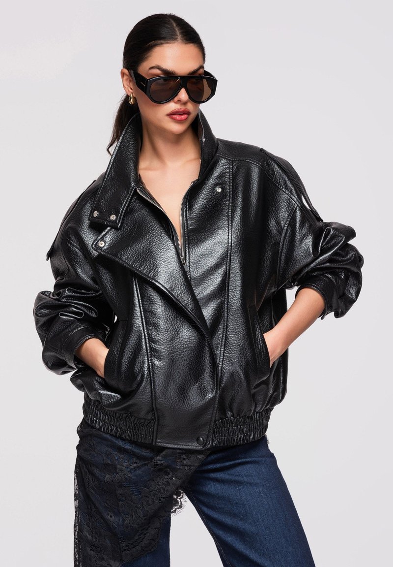 Mujer con chaqueta de cuero negra de gran tamaño, gafas de sol oscuras y vaqueros, posando con las manos en los bolsillos frente a un fondo liso.