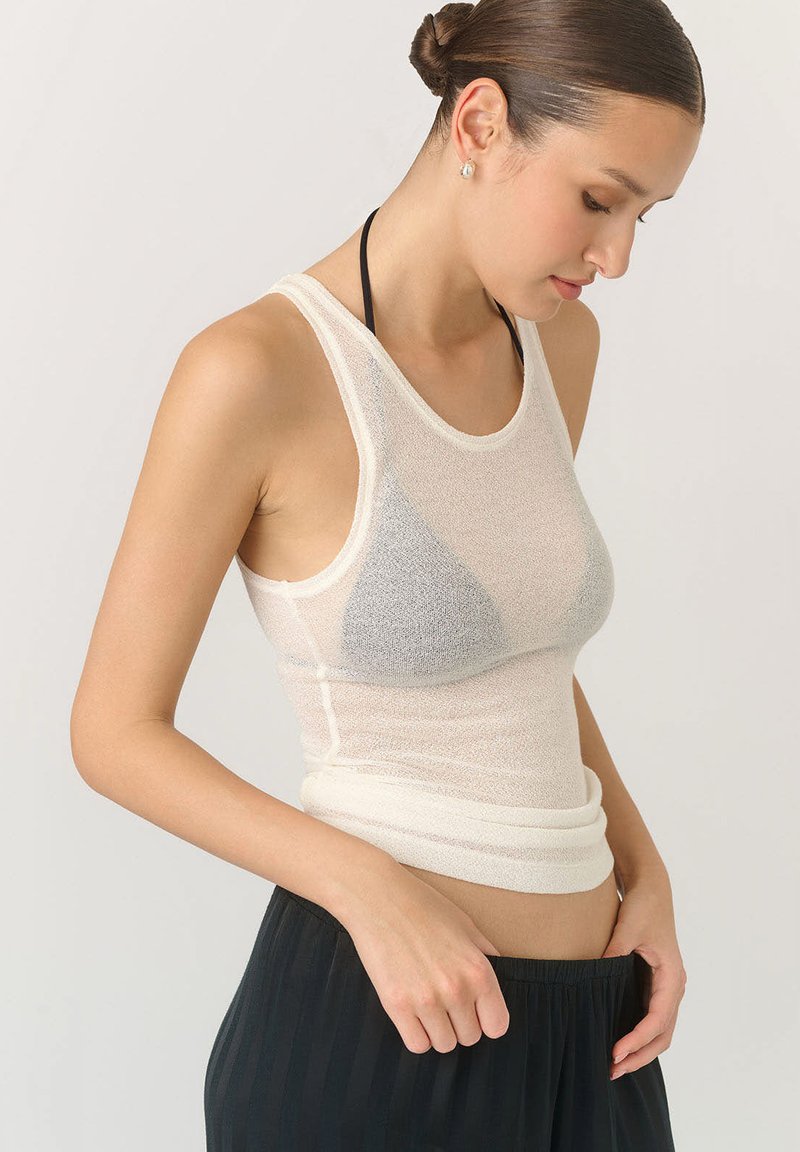 Biała, dopasowana koszulka na ramiączkach z przejrzystą teksturą, z lekkiego, przepuszczającego powietrze materiału oraz okrągłym dekoltem. Nosi się ją z ciemnymi, zwiewnymi spodniami.