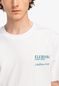 Bílý bavlněný tričko s kulatým výstřihem, se tyrkysovým textem "ELEMENT 75000 PARIS" a "L'Addition SVP" na levé části hrudi.