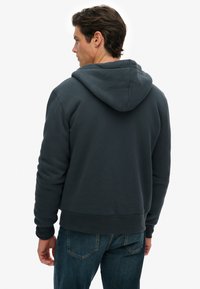 Man in een donkergrijze capuchontrui en blauwe jeans, staand met zijn rug naar een witte achtergrond.