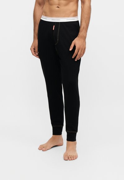 Homme portant un pantalon noir ajusté avec une ceinture élastique blanche marquée "DSQUARED2,", debout pieds nus devant un fond clair uni.