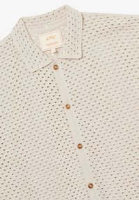 Chemise en tricot crochet beige avec un col pointu et quatre boutons ronds marron sur le devant.