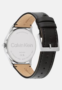 Zegarek Calvin Klein z stalową kopertą, czarnym skórzanym paskiem i srebrnymi elementami. Posiada gładką matową teksturę i odporność na wodę.