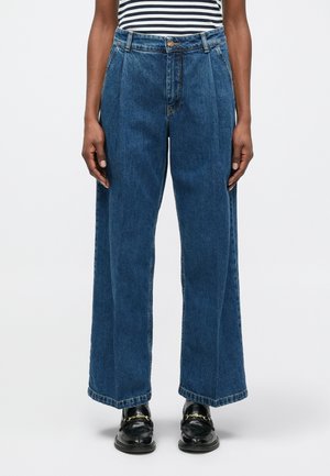 Osoba oblečená v vysokých, širokých modrých džínsoch, skombinovaných s čiernymi mokasínami so zlatým reťazovým detailom a pruhovaným topom.