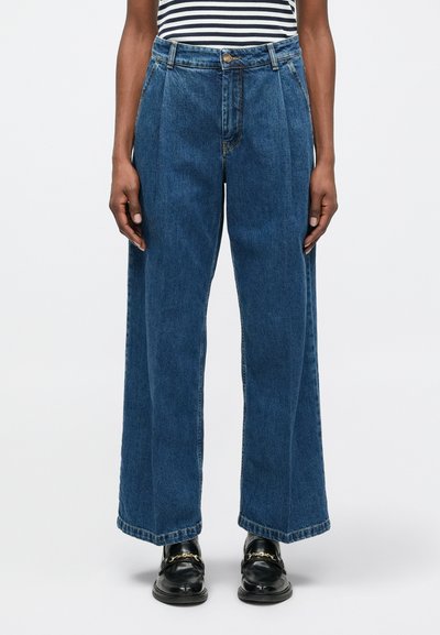 Persona que lleva jeans azules de talle alto y pierna ancha, combinados con mocasines negros con detalle de cadena dorada y una camiseta a rayas.