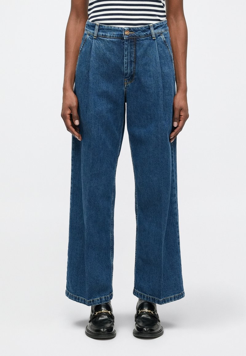 Person iført høy midje og vide blå jeans, kombinert med svarte loafers med gullkjede-detalj og en stripete topp.