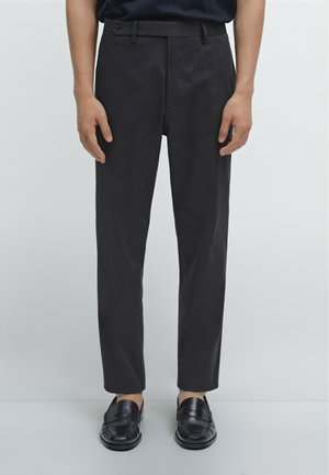 Pantaloni neri slim-fit con parte anteriore piatta, dotati di tasche laterali e chiusura a un bottone. Indossati con mocassini neri. Texture liscia.