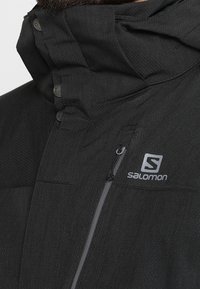 Veste d'extérieur Salomon noire avec poche à fermeture éclair, col haut boutonné et tissu texturé visible sur la partie supérieure de la poitrine et du cou.