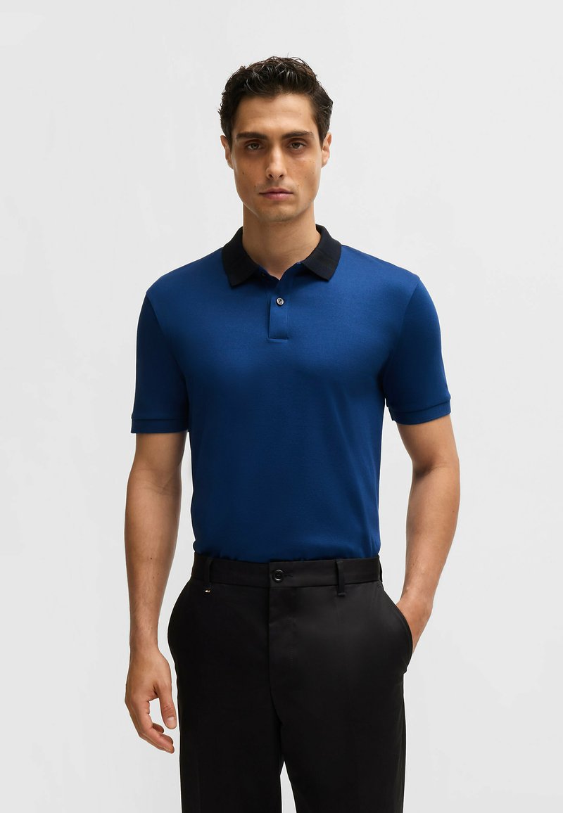 Polo shirt bleu à manches courtes et col noir, avec une patte de boutonnage. Assorti à un pantalon noir, le tissu semble lisse.