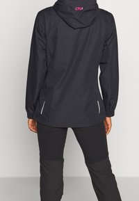 Veste noire imperméable avec capuche, accents blancs, et logo CMP en rose au dos. Matière légère, poignets élastiques et coupe décontractée.