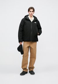 Zwarte waterdichte jas met capuchon en ritssluiting, borstlogo, gecombineerd met kaki broek en zwarte pet, staande op een effen achtergrond.