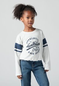 Nuori tyttö, jolla on kiharainen tukka, pukeutuneena valkoiseen svetariin, jossa on sinisiä raitoja, sekä farkkuihin, seisoo yksinkertaista harmaata taustaa vasten.