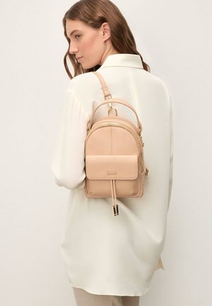 Mały beżowy plecak ze skóry o zaokrąglonym kształcie, z przednią kieszenią, złotymi ozdobami, górnym uchwytem oraz regulowanym paskiem.
