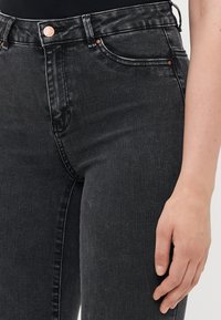 Nærbilde av en person som har på seg høy midje sorte denimjeans med kobberniter, og en synlig høyrehånd som hviler ved siden av låret.