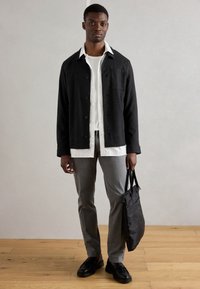 Veste noire à boutons, chemise rayée blanche en dessous, pantalon gris, chaussures noires, tenant un sac fourre-tout noir, debout sur un sol en bois.