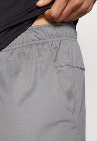 Γκρι σορτς με ελαστική μέση, υφασμάτινη υφή και λεπτομέρεια πλαϊνής τσέπης. Το εσωτερικό δείχνει δέρμα κάτω από ένα μαύρο πουκάμισο.