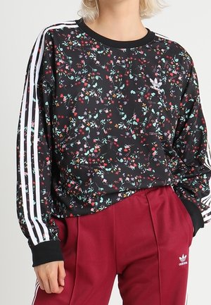 Czarna, kwiatowa, długorękawowa bluzka Adidas z białymi paskami na rękawach, zestawiona z czerwonymi spodniami Adidas z białym logo i paskami.