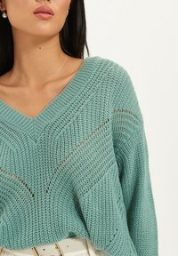 Pull en maille à col en V turquoise, avec un détail en maille ajourée et une coupe ample. Un motif texturé traverse le tissu, ajoutant de l'intérêt visuel.
