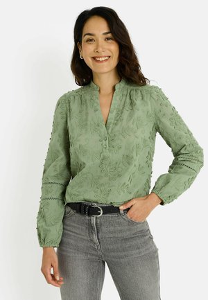 Blusa abotonada verde claro con bordado floral, mangas largas y cuello mao, combinada con jeans grises y cinturón negro.