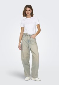 Camiseta blanca con patrón de puntos negros y mangas cortas, combinada con jeans holgados de color azul claro y zapatillas blancas, de pie en un fondo neutro.