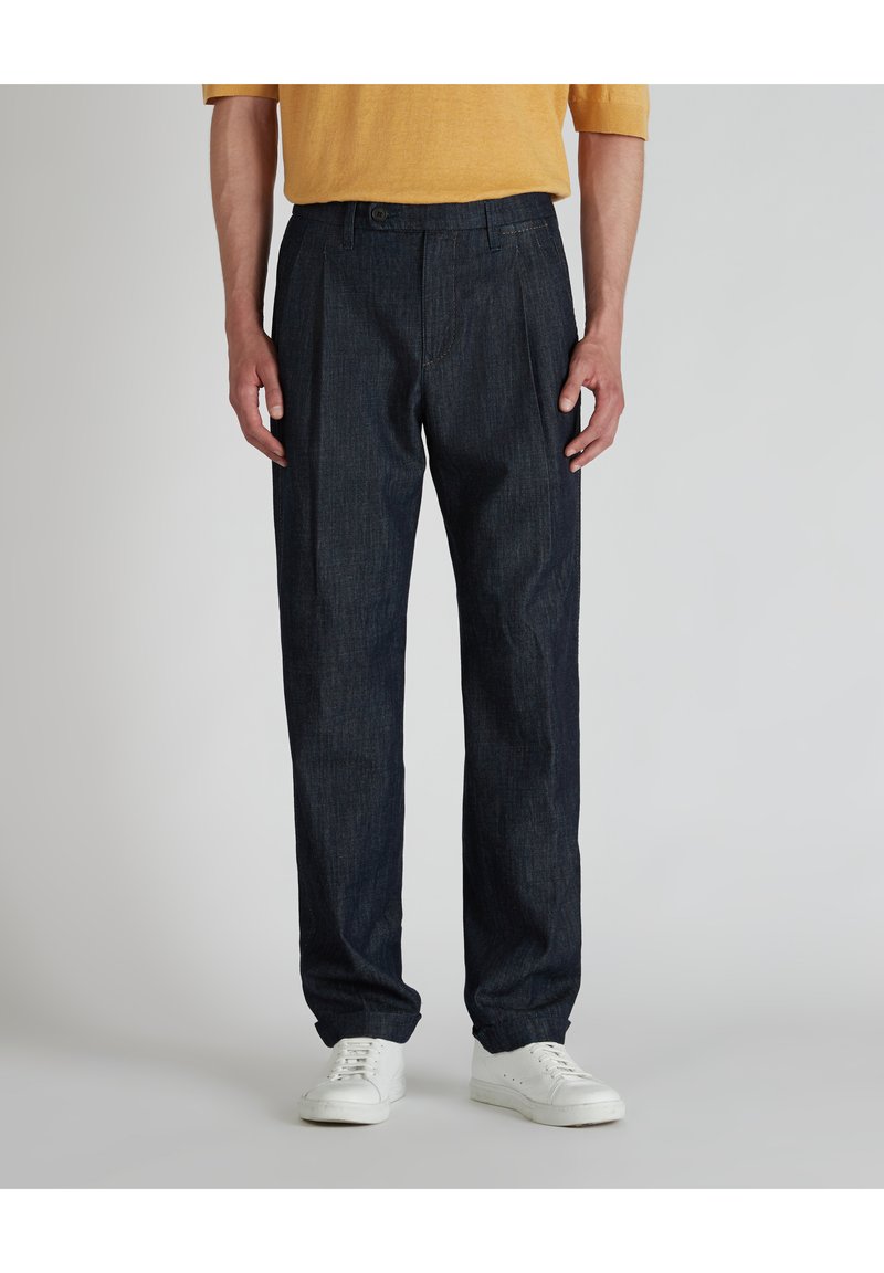 Pantaloni a pieghe blu scuro in denim, con chiusura a bottone e tasche laterali, abbinati a sneakers bianche.