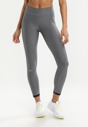 Femme portant des leggings de sport gris moulants et des chaussures de course blanches, se tenant devant un fond blanc.