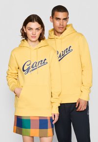 Gul genser med en stor blå "Gant" logo, lommer foran og snøring på hetten. Modellen parrer den med et flerfarget rutete miniskjørt.