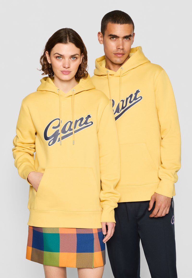 Gul genser med en stor blå "Gant" logo, lommer foran og snøring på hetten. Modellen parrer den med et flerfarget rutete miniskjørt.