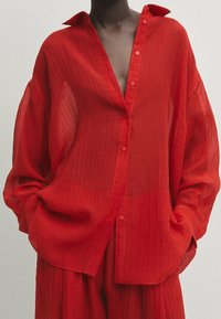 Chemise rouge transparente à boutons avec col cranté et manches longues, présentant un tissu texturé et une coupe ample.