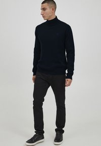 Pulover din lână de culoare bleumarin, fabricat dintr-un material tricotat moale, având mâneci lungi și margine ribbată. Asortat cu pantaloni închiși la culoare, ajustați pe corp, și adidași.