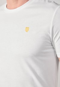 T-shirt en coton blanc avec un col rond et des manches courtes. Détail notable : un petit logo de bouclier brodé jaune sur la poitrine.