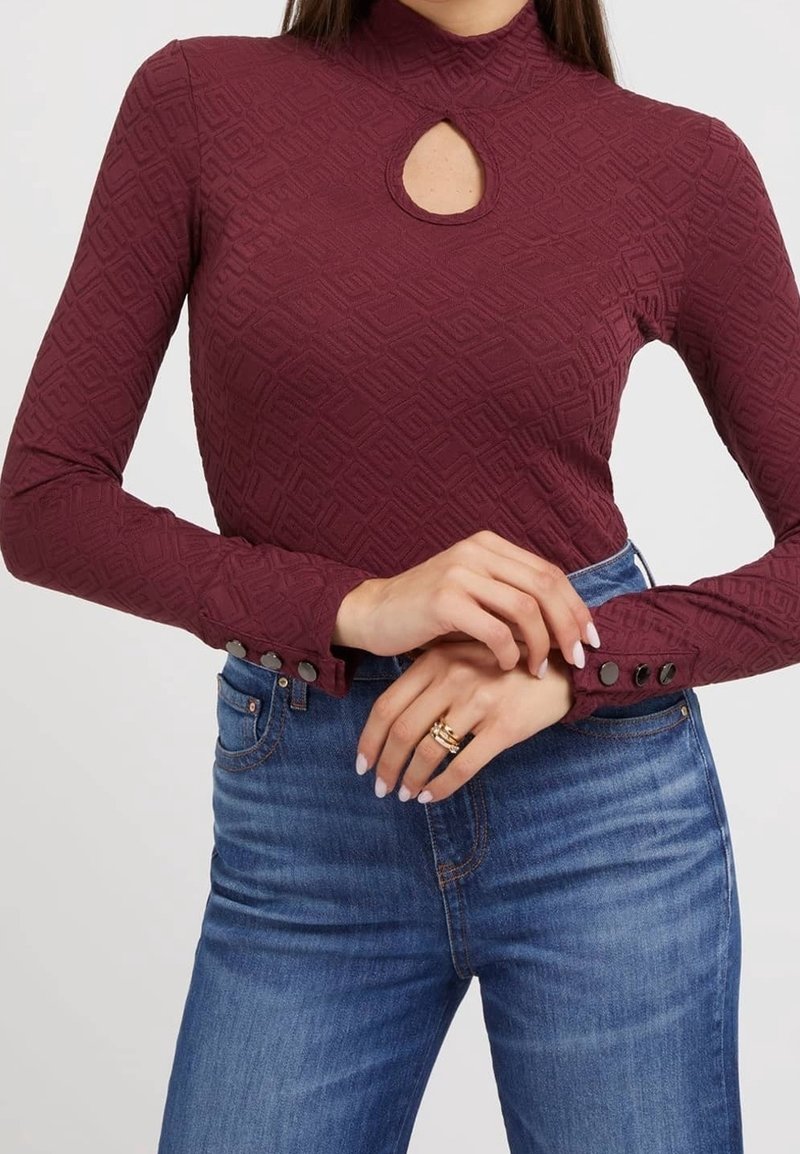 Bordeaux langearmige top met sleutelgat-halslijn, textuur geometrisch patroon en knoopaccenten bij de mouwen, gedragen met blauwe jeans.