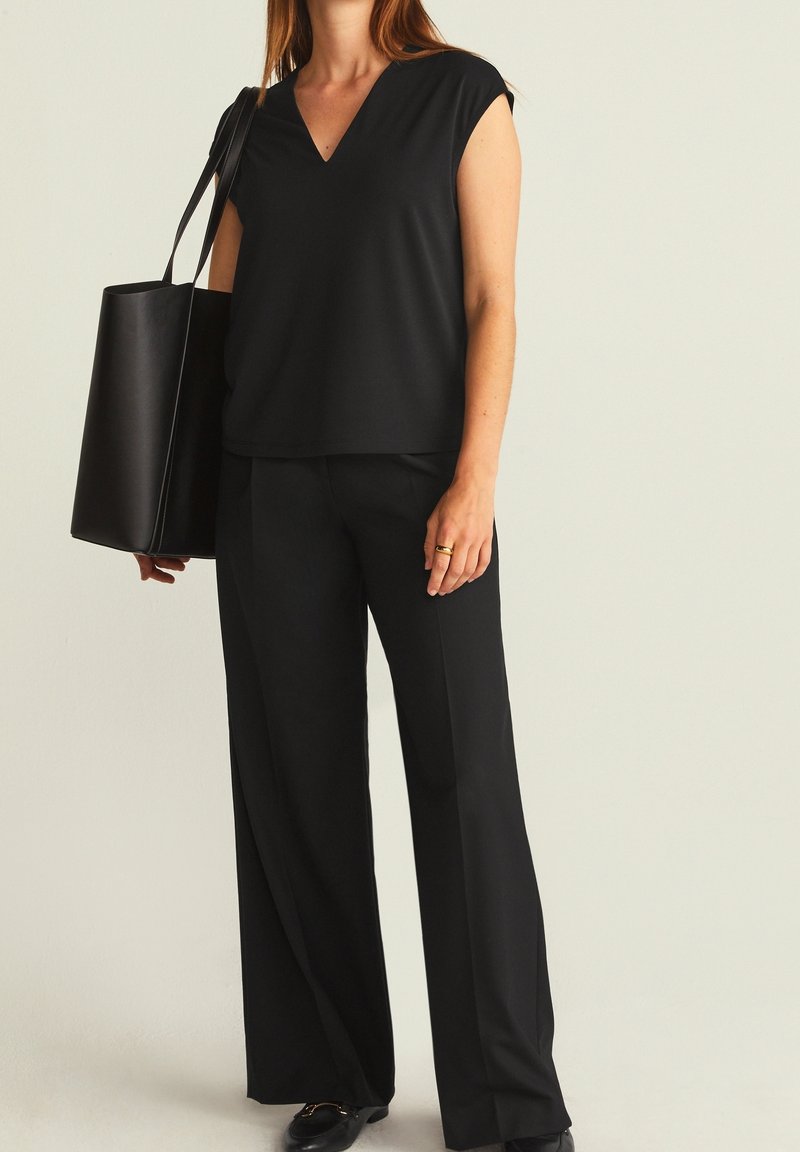 Femme portant un pantalon large noir et un haut sans manches à col en V, avec un grand sac cabas noir à l'épaule.