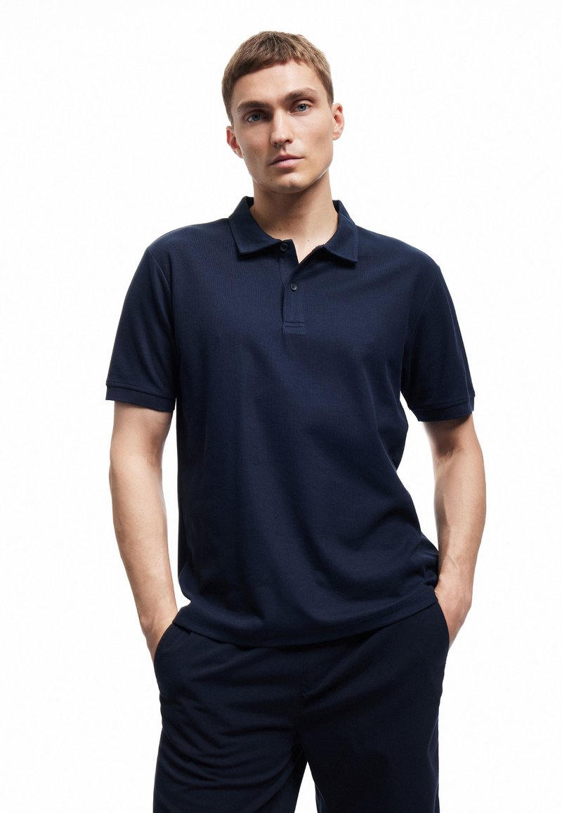 Marineblauw poloshirt met een textuurstof, korte mouwen en een kraag met twee knopen. Draagt met bijpassende marineblauwe broek.