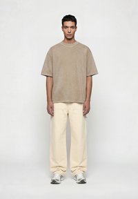 Νεαρός άνδρας που στέκεται πρόσωπο, φορώντας μια υπερμεγέθη μπεζ μπλούζα, κρεμ παντελόνια με φαρδιά πόδια και λευκά αθλητικά παπούτσια σε απλό φόντο.