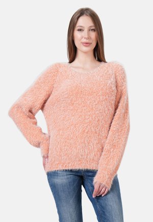 Flauschiger orangefarbener Pullover mit langen Ärmeln und weitem Ausschnitt, der eine strukturierte Oberfläche und einen lockeren Sitz hat. Getragen mit blauen Jeans.