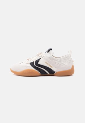 Baskets basses blanches avec semelle en caoutchouc beige, motif ondulé noir sur le côté, col en maille autour de la cheville, et texte de la marque « Steve Madden » sur la semelle et le côté.